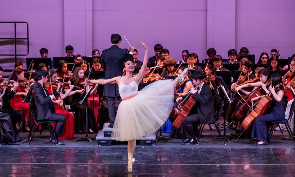 A ballet dancer in a knee-length tulle dress on pointe in front of a string orchestra.