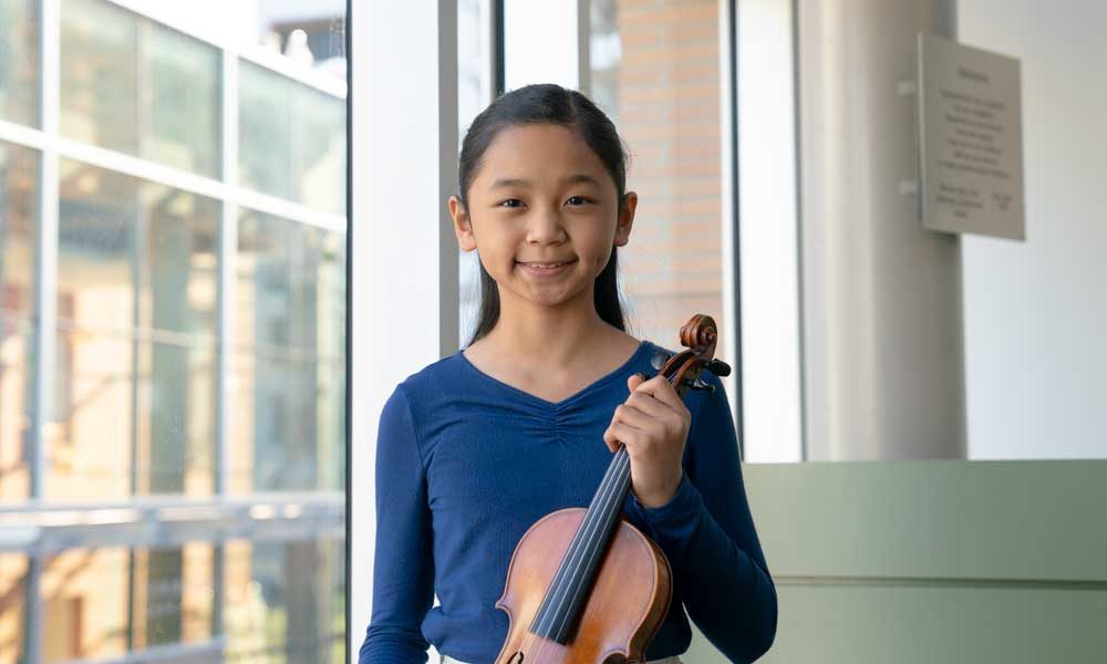 An elementary school aged student holding a violin and smiling in front of a window.