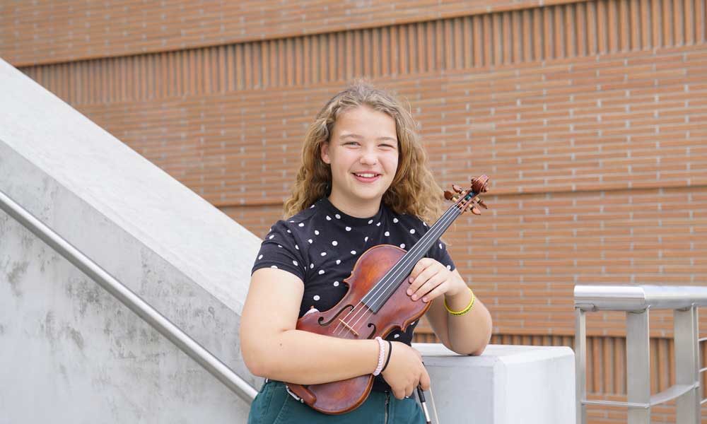 A young student holding a viola and leaning on a railing, smiling.