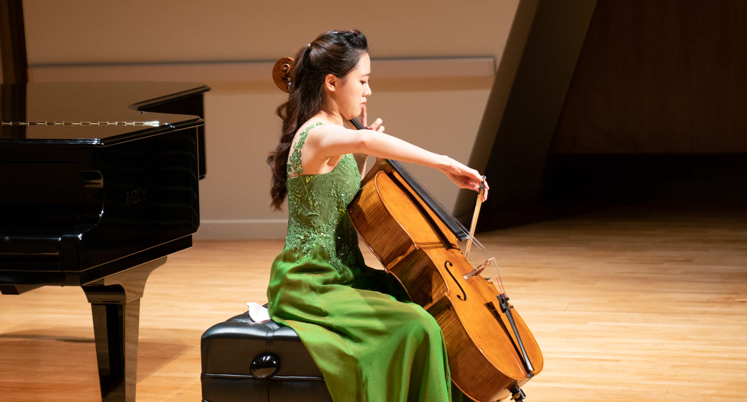 Sanga Yang in a dress playing cello in front of a piano.