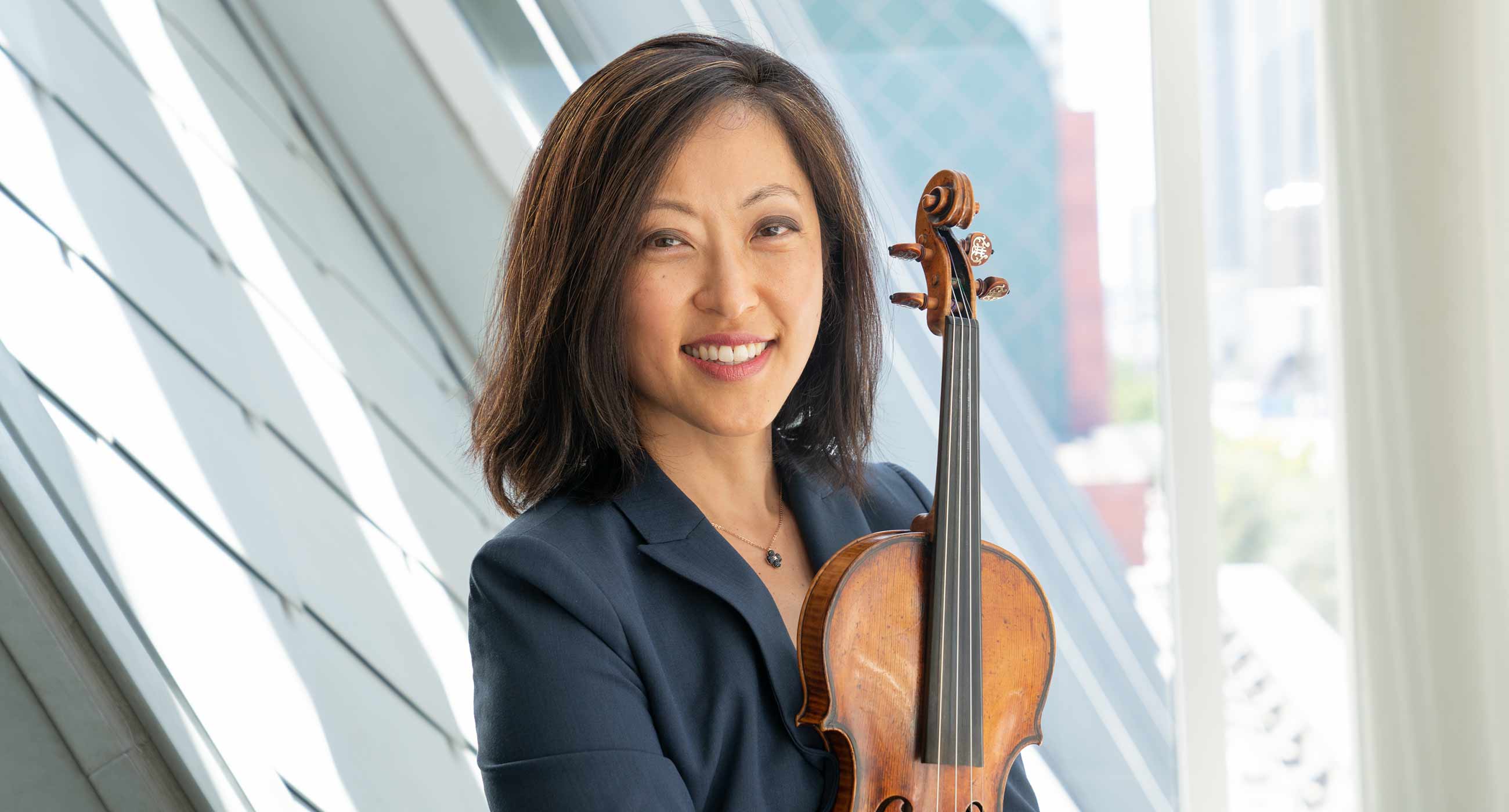 Joan Kwuon wearing a blazer, holding a violin, and smiling in front of a window.