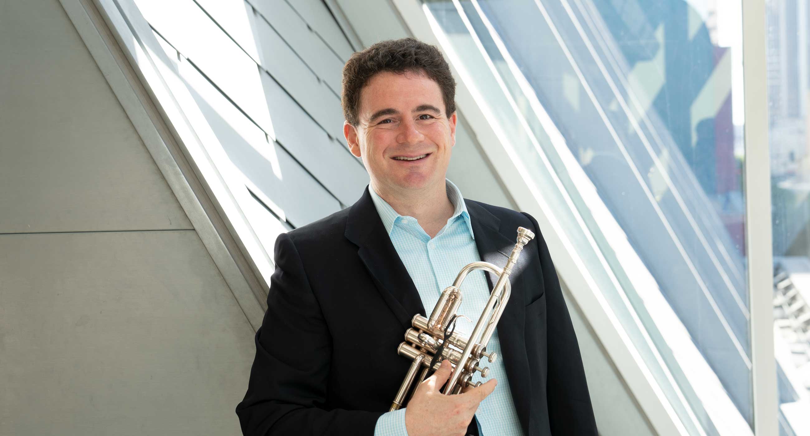 Mike Zonshine wearing a collared shirt and a suit jacket holding a trumpet in front of a window.