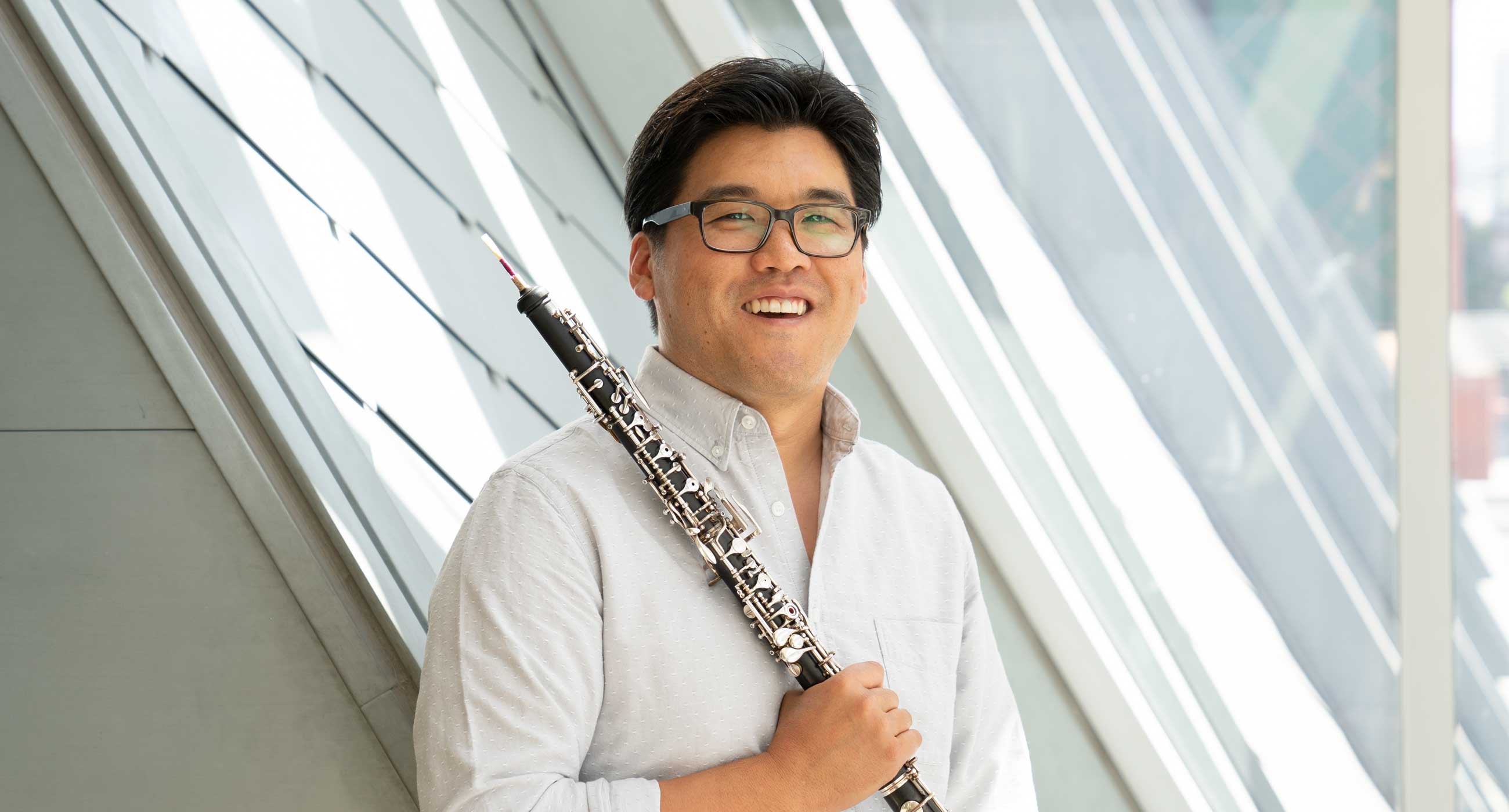 Ted Sugata holding an oboe and smiling in front of a window. He is wearing glasses.