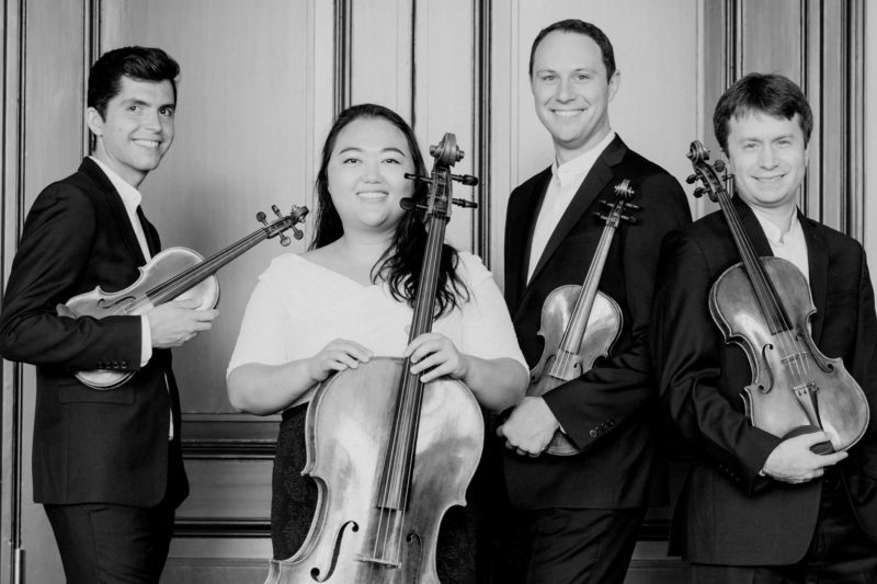 The Calidore Quartet holding their instruments and smiling in front of a fancy set of doors.