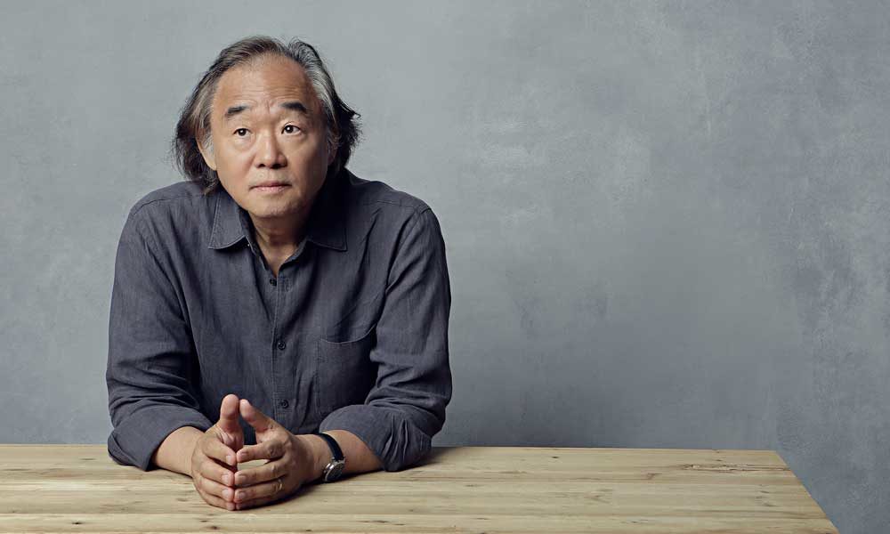 Kun-Woo Paik leaning on a table wearing a collared shirt and a watch.