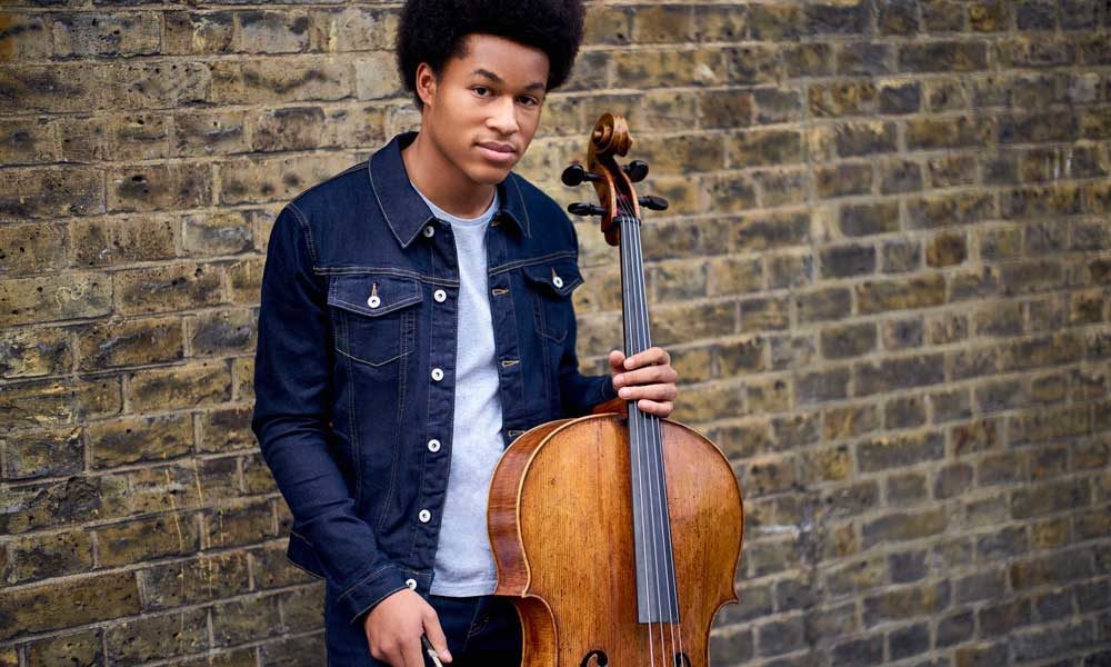 Sheku Kanneh-Mason holding a cello and leaning against a wall.