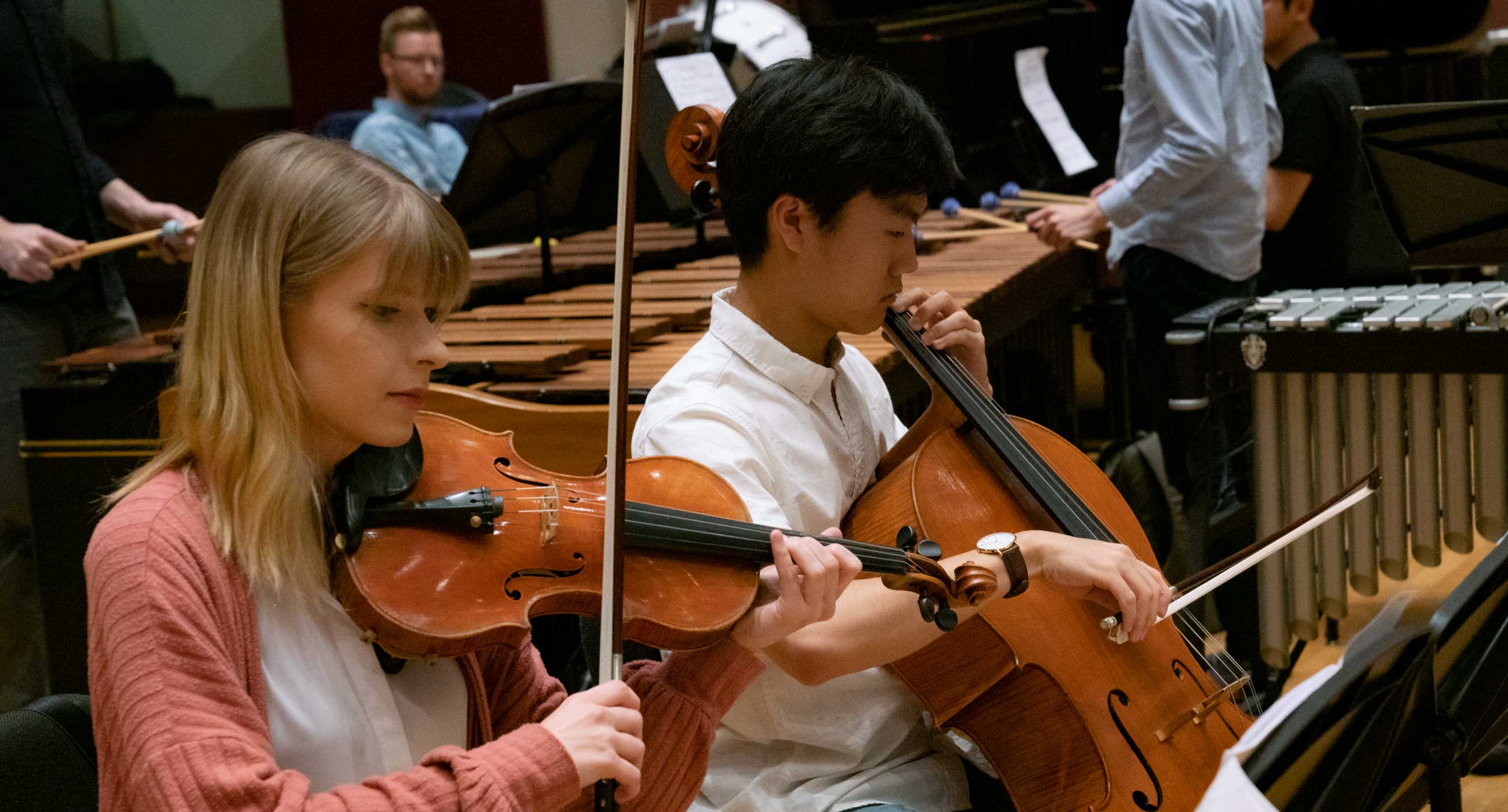 Two students, one playing violin and one playing cello, in front of several marimbas.