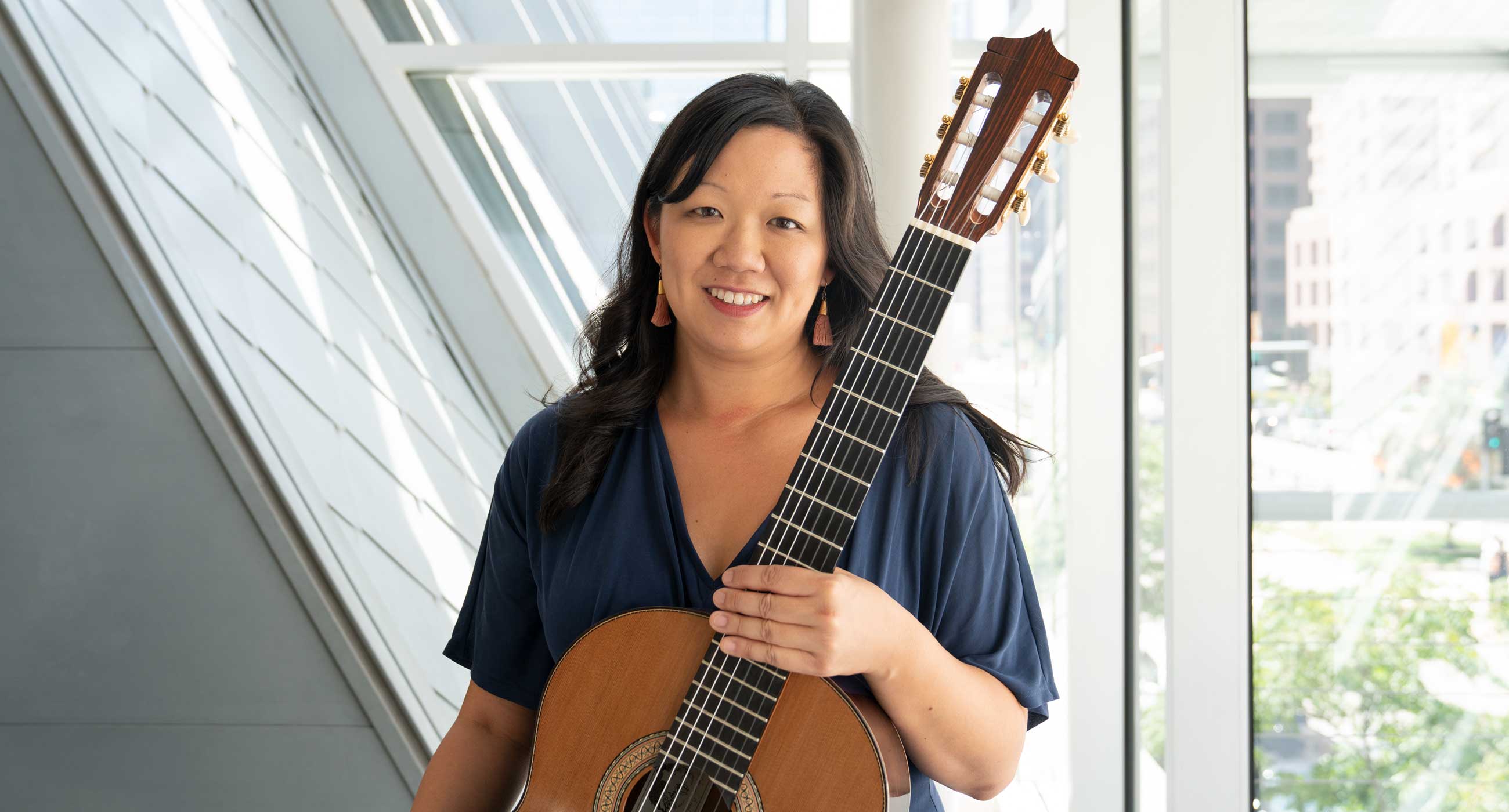 Connie Sheu holding a classical guitar and smiling in front of a window. She is wearing tassel earrings.