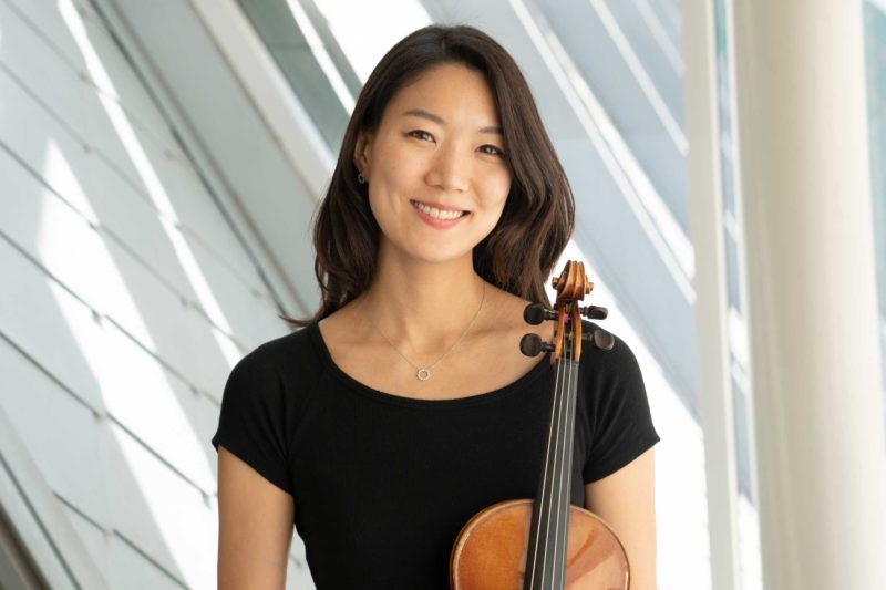 Joanna Lee holding a violin and smiling in front of a window.