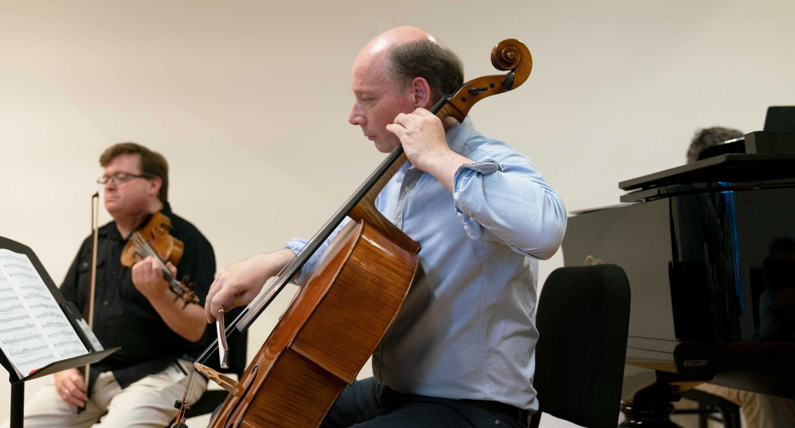 Clive Greensmith playing cello next to a piano with violinist in background