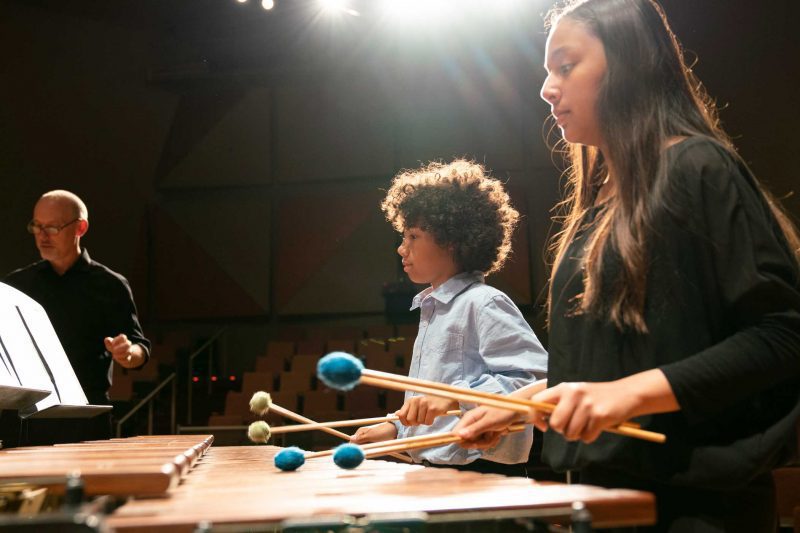 Girl and boy with playing marimba with mallets in hands on stage, teacher Ken McGrath in background