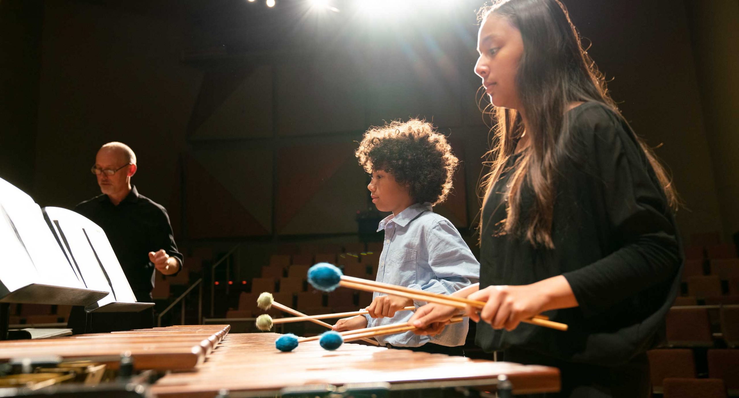 Girl and boy with playing marimba with mallets in hands on stage, teacher Ken McGrath in background