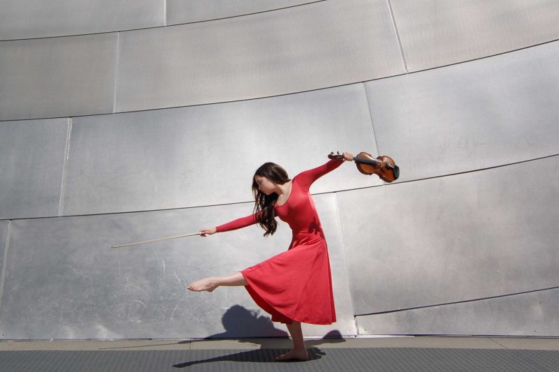 Holly Lacey wearing a red long sleeve dress posing with a violin in one hand, standing in front of a silver metal wall