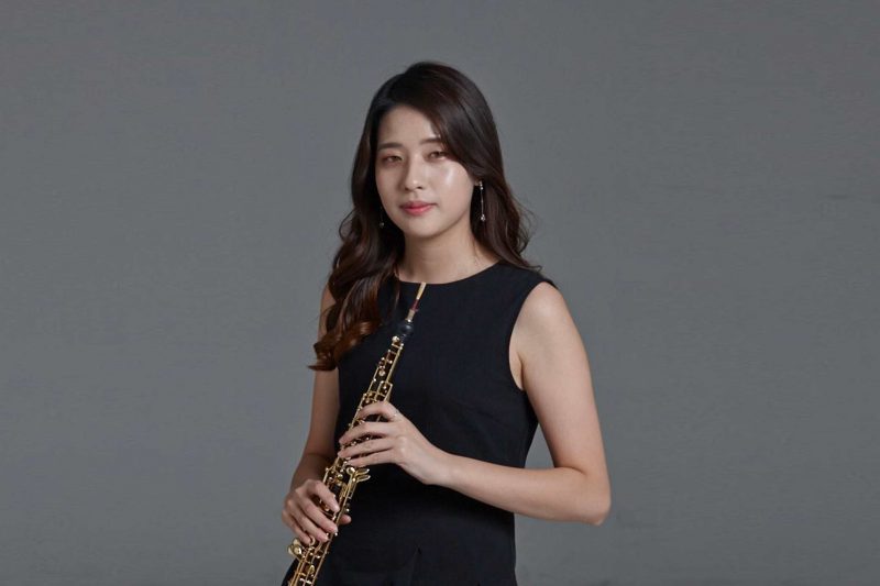 Korean woman with long black hair holding a oboe wearing a black dress against a grey background