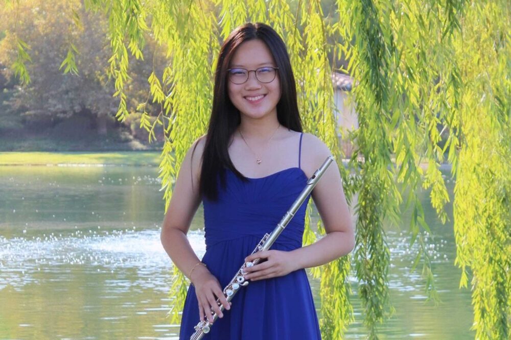 Celine Chen wearing a blue dress holding a flute, standing in front of a pond with tree branches with green leaves