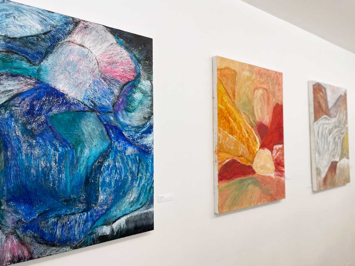 Three paintings on a wall from a diagonal angle. Closest painting is of grey rocks and blue water. Middle painting is of orange and red rocks. Furthest painting is of grey and brown wavy shapes.